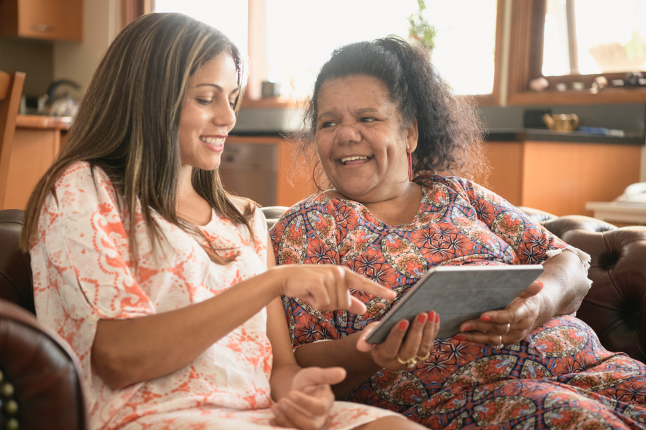 Image of smiling elderly woman and younger woman on a couch pointing at a tablet devices screen.