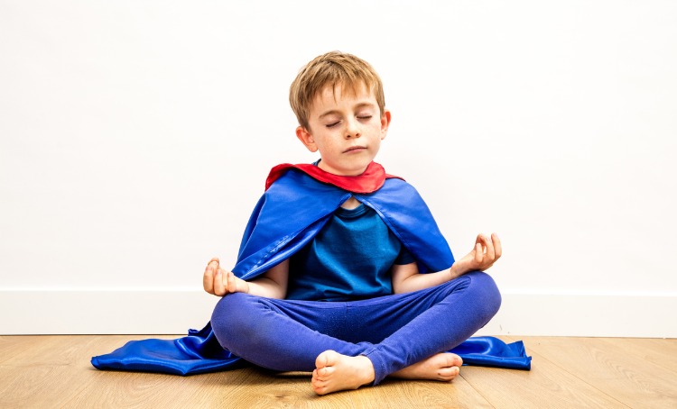 A blonde haired boy wears a superhero cape and outfit in blue and red. He is sitting in a Yoga pose, his feet are crossed, hands on his knees with thumbs to fingers and his eyes are closed.