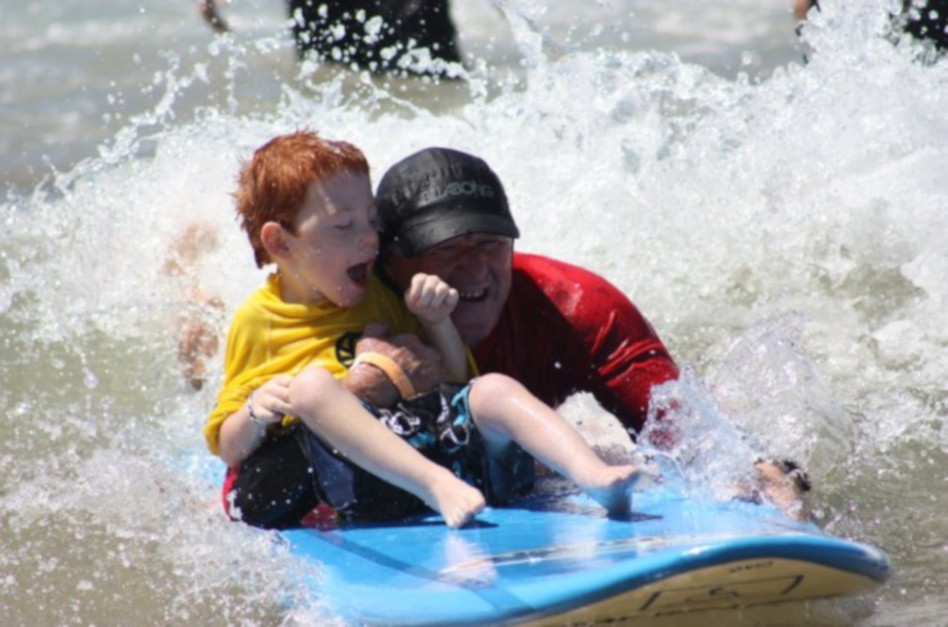 An image of a young boy on a surfboard in the ocean with a volunteer.