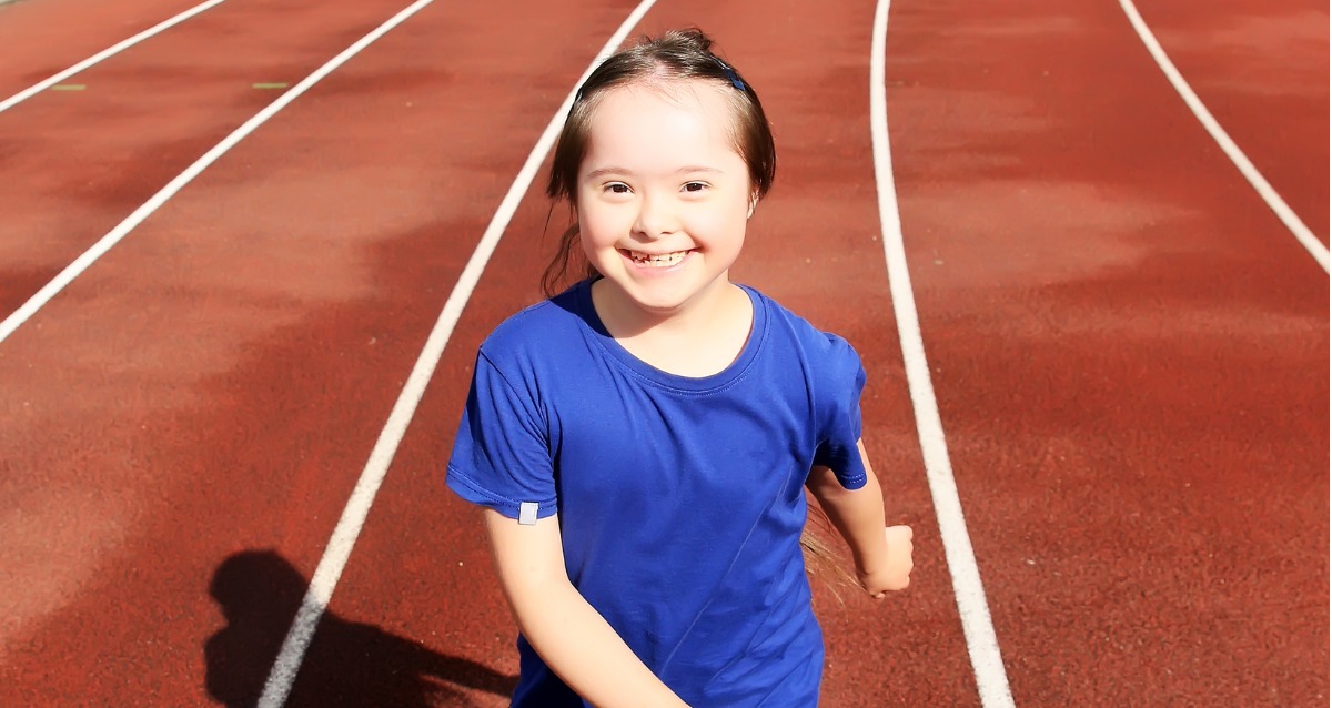 A photo of a young girl with down syndrome smiling at the camera. She is standing on an athletics track.