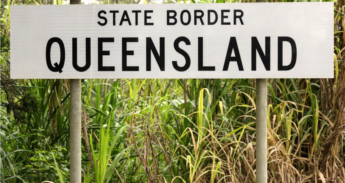 A photo of a State Border Sign, with the writing "State Border Queensland" in black on the white sign. 