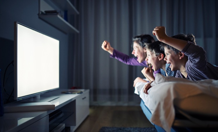 3 kids watching tv and with raised arms like cheering picture. The room is semi-darkened and they are lit up by the TV. They are laying on a couch or bed.