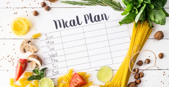 A printed piece of paper with the heading "Meal Plan" has days of the week and columns to place meals. The paper is surrounded by foods like pasta, herbs, vegetables, fruits, nut and spices,