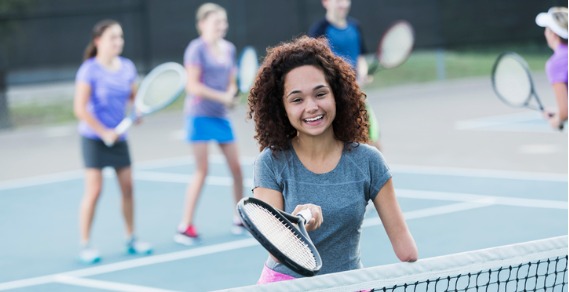 An Image of a young girl with disabilty, standing at a tennis net, holding a tennis racquet