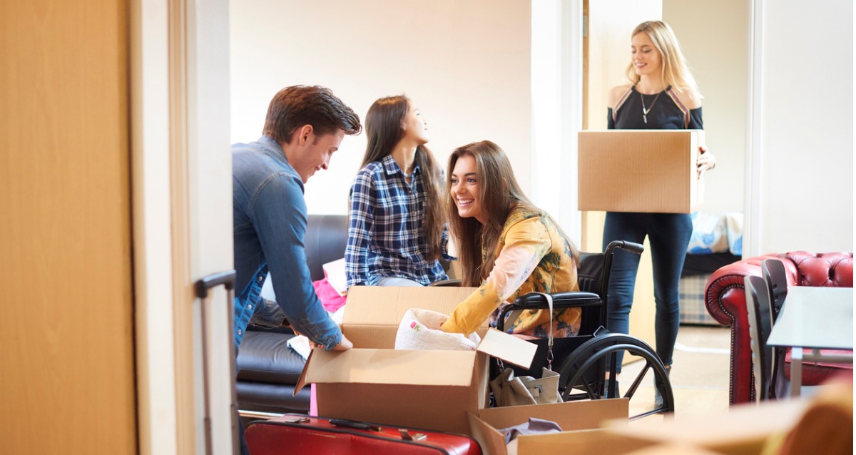 A group of University students are moving into a shared flat. They are surrounded by boxes. One student is in a manual wheelchair.