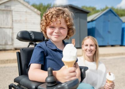 Young boy in wheelchair holding an ice-cream. Beside him is a woman holding an ice-cream. In the background are beach huts.