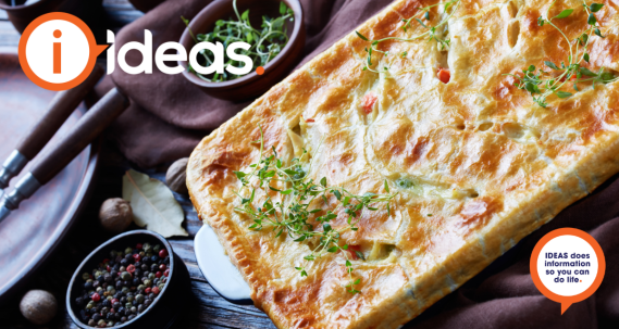 A table set with a pie dish, cutlery, crockery and a nutmeg, bayleaf and peppercorns. The pie is golden with vegetables showing through some slits, it is garnished with herbs such as thyme.