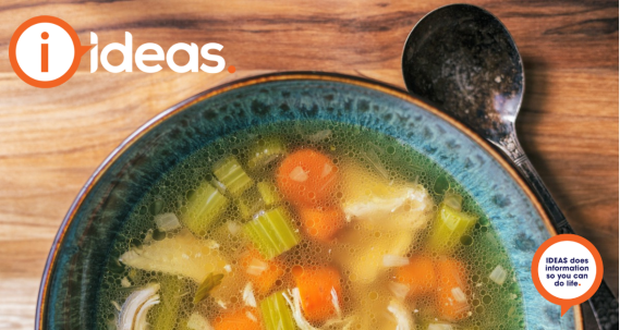 A blue bowl with a brown edge sits on a timber table. Beside it is a spoon. In the bowl is broth, celery, chicken, carrot and onion. A bowl of Chicken soup.