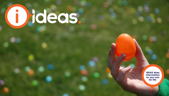 A small hand holds up an orange coloured plastic easter egg. In the background are blurred easter eggs on green.