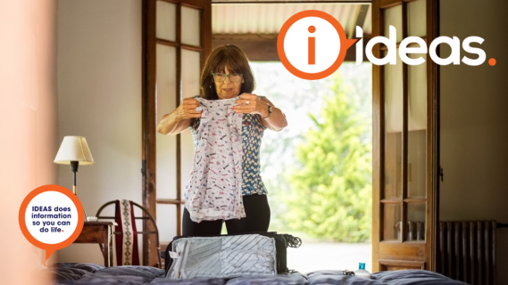 A woman holds an item of clothing to pack in a suitacse which is on the bed in front of her.