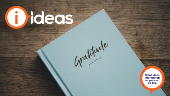 A pale blue hard bound book, with the words "gratitude journal" written on the front cover in a mixture of cursive and plain text rests on a timber tabletop.