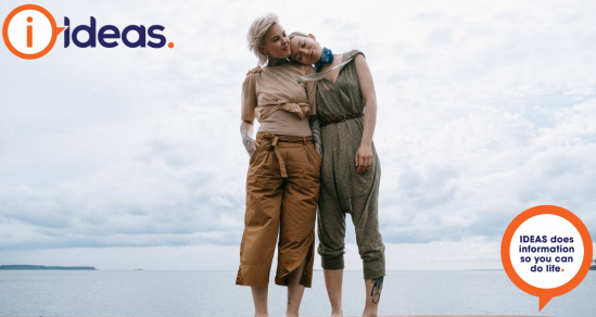 Two women standing together supporting one another. In the background is water and a cloudy sky.