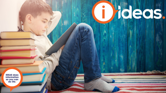 A young boy sites with a book on his lap reading, Beside him is a pile of books.