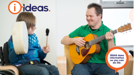 Two people. One is holding a drum, and one a guitar, they are making music and smiling. 