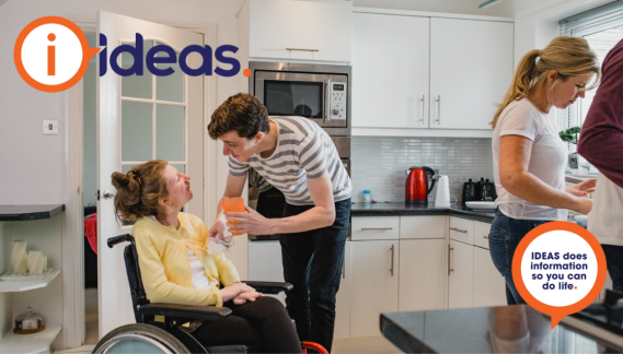 A young male helps a girl to take a drink. She is in a wheelchair. They are in the kitchen with 2 other adults. 
