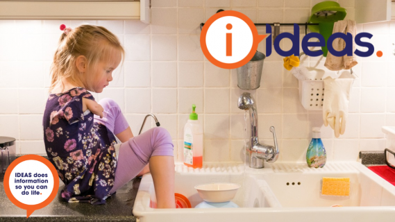 A young girl sits on the kitchen bench beside the sink. She washes the dishes with her feet. 