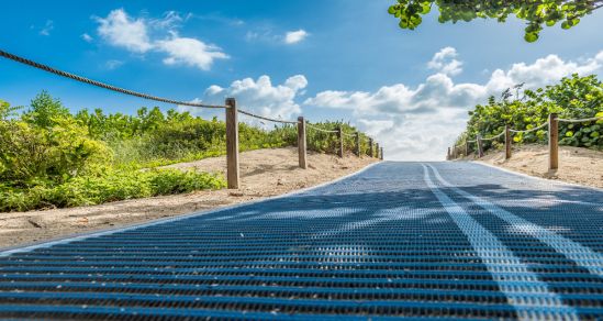 A photograph of a beach pathway with accessible matting. In the background is a blue sky with clouds.