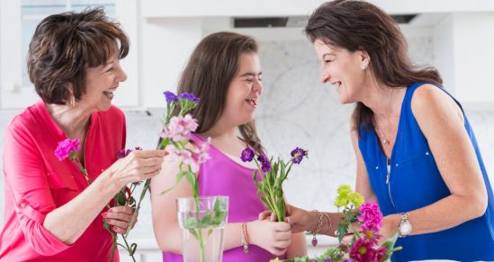 An image of three women of different generations. The youngest is a woman with disability. They are laughing and arranging flowers.