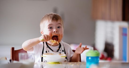 A toddler with disability is sitting in a high chair and feeding himself spaghetti. His cheeks are covered in food and his mouth is open wide to put a spoonful in.