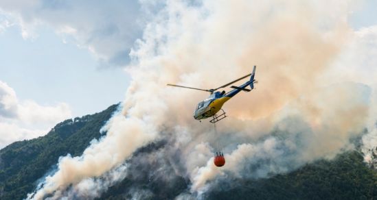 Helicopter with a bucket attached flyer over hills that are on fire