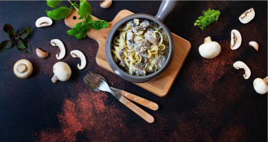 A concept photo of mushroom stroganoff, a dish with mushrooms and pasta, aongside is a knife and spoon. Around the bowl are mushrooms, basil, herbs and paprika.