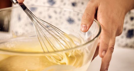 Image of a womans hands, holding a whisk, mixing a batter in a bowl.