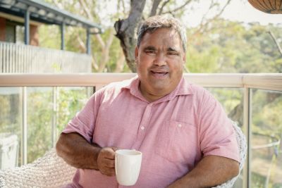 Image of smiling elderly Aboriginal man sitting outside at a table with a hot drink.