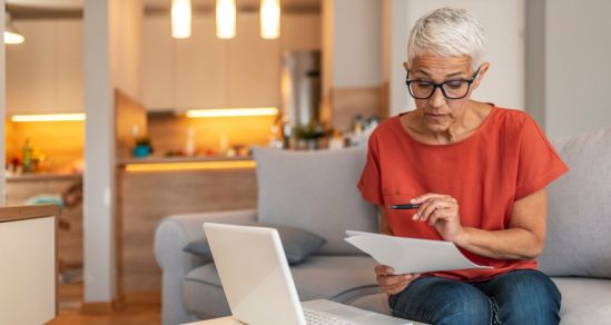 A woman wearing glasses and a red shirt, holds a pen and is checking some paperwork. In front of her is a white laptop.