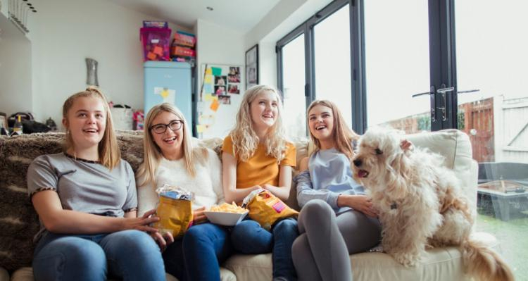Four teenage girls, one with disability on a lounge. The girls have snack food and a labradoodle sits at one end. They are laughing, smiling and relaxed.