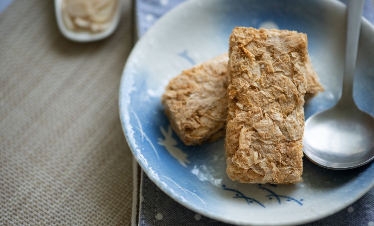 Image of whole grain wheat biscuits cereal. Two are placed in a bowl that is clue and white, a spoon rests beside the, The tablecloth is tan colour.