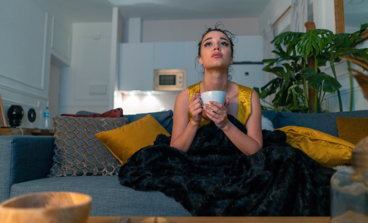 A young woman holding a cup of warm drink is swathed in a blanket, sitting on the couch. Behind her pillows are propped up and she watches the TV which is out of the picture.