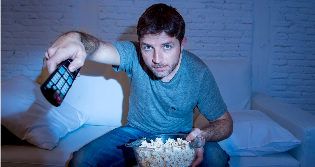 A man holds a remote pointed to the TV, on his lap he has a bowl of popcorn. He is sitting on a lounge in a semi-darkened room. The light from the TV reflects from his face.