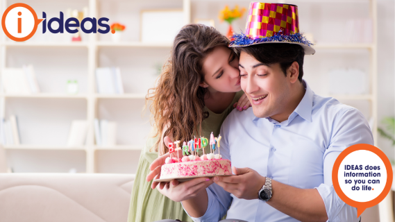 Young man celebrating his birthday with a loved one. He is holding a cake and wearing a party hat.