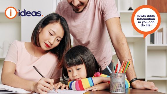 a woman and small child sitting and writing with pencil and paper. a man stands behind them overllooking. 