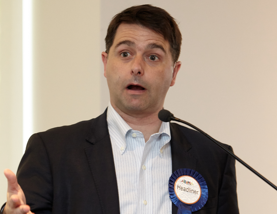 Image of Alastair McEwin speaking and wearing a blue show ribbon labelled "Headliner"