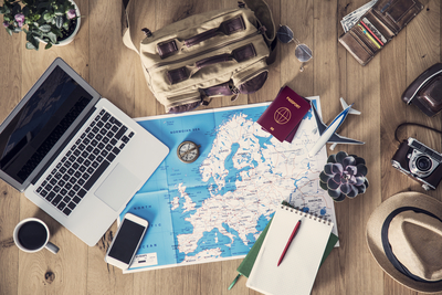 Image of maps, notebook and passport on a desktop with laptop, phone and coffee mug.
