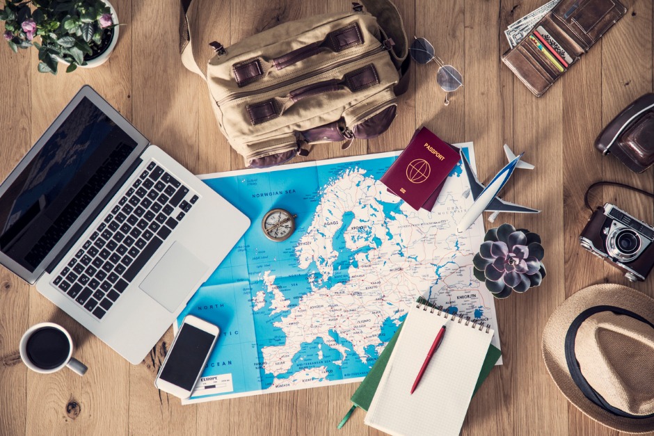 Image of maps, notebook and passport on a desktop with laptop, phone and coffee mug.