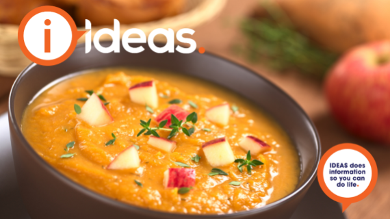 A black bowl containing a orange coloured soup, with diced red apple and thyme as a garnish.
