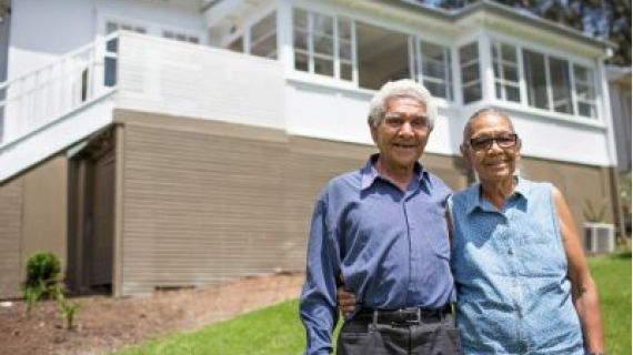 Image of elderly couple smiling with their arms around each other and standing in front of a house.