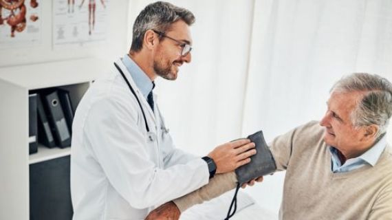 A Doctor wearing white coat and glasses has a stethoscope around his neck. He is holding a blood pressure monitor and wrapping the band around a patients arm in preparation to take blood pressure reading.