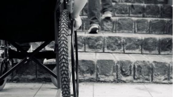 Black and white image of wheelchair in front of a flight of stairs, being unable to go up them.