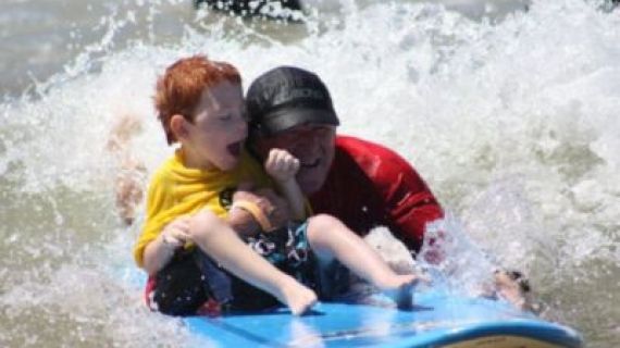 An image of a young boy on a surfboard in the ocean with a volunteer.