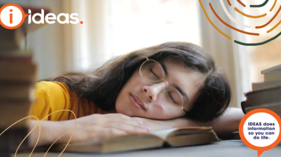tired woman with glasses sleeping with face on open book. IDEAS logo is in upper left corner.