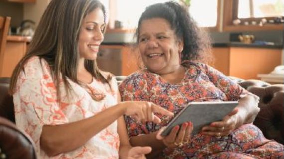 Image of smiling elderly woman and younger woman on a couch pointing at a tablet devices screen.