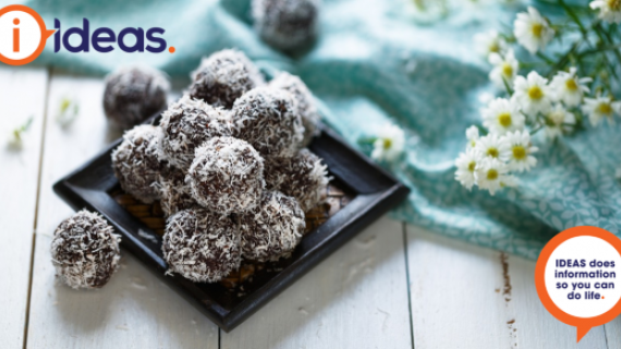 An image of bliss balls stacked on a black plate. They are coated in coconut. A blue floral tablecloth and some flowers are on the right of the photo