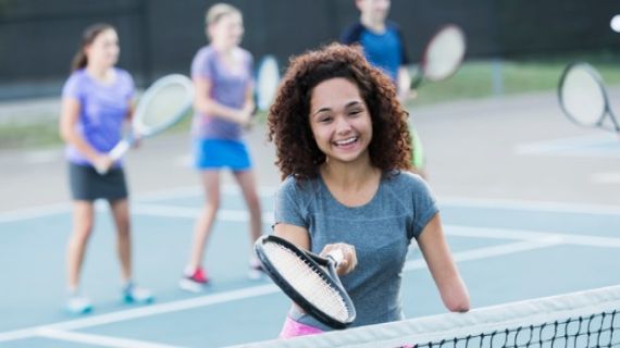 An Image of a young girl with disabilty, standing at a tennis net, holding a tennis racquet