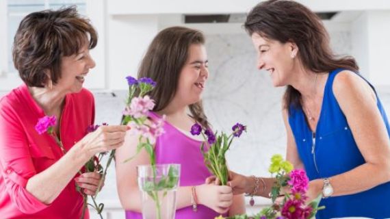 An image of three women of different generations. The youngest is a woman with disability. They are laughing and arranging flowers.