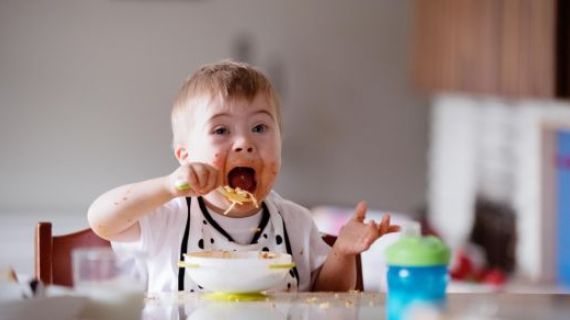 A toddler with disability is sitting in a high chair and feeding himself spaghetti. His cheeks are covered in food and his mouth is open wide to put a spoonful in.