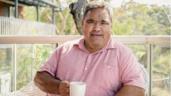 Image of smiling elderly Aboriginal man sitting outside at a table with a hot drink.
