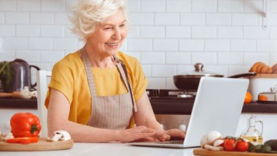 An image of an older woman in a kitchen looking at a laptop. She is wearing an apron and is surrounded by vegetables and cooking utensils